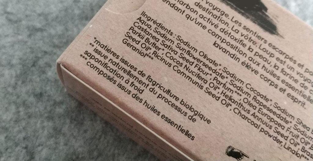 Gros plan d'un emballage carton kraft de shampooing solide artisanal tenu dans une main, étiquette avec liste d'ingrédients INCI en français visible, fond neutre flouté, lumière naturelle